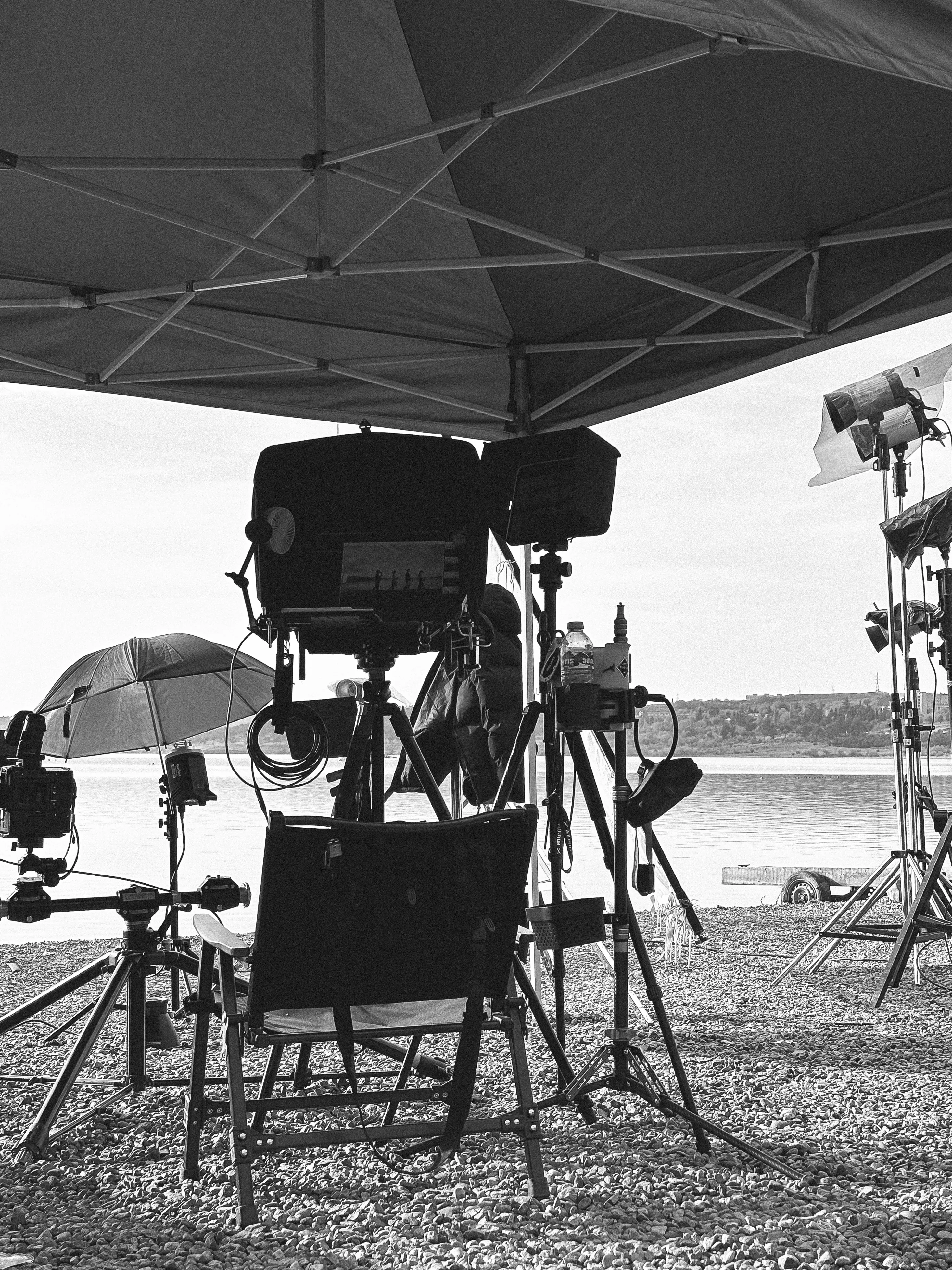 Black and white photo of film production equipment and a director's chair on a rocky shore by a calm body of water.